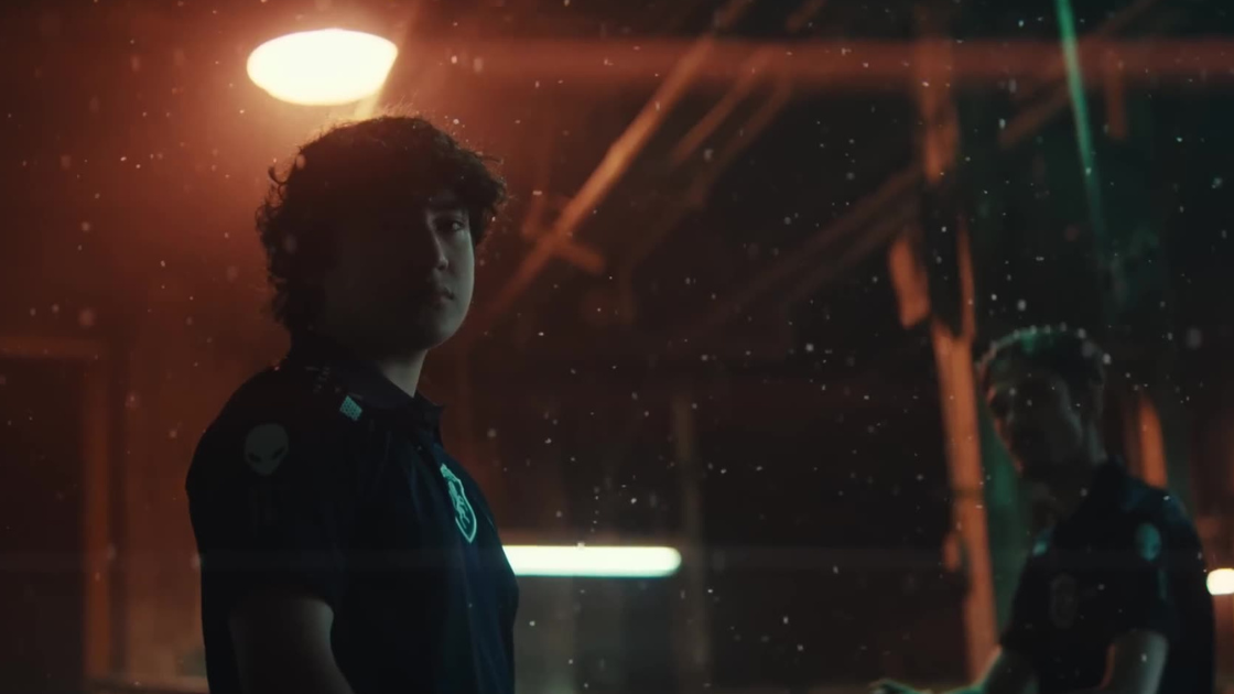 A player stands still under a warm overhead light, surrounded by drifting particles. His focused stare contrasts the shadowed figure in the background, evoking tension, quiet confidence, and a sense of readiness in a charged, cinematic space.
