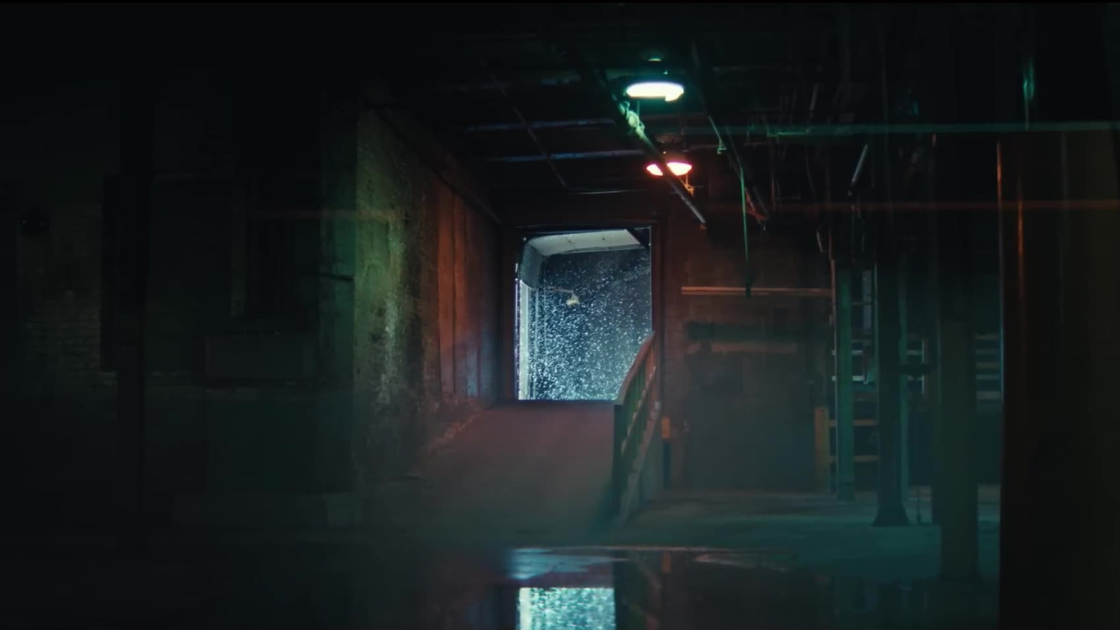 A dimly lit loading dock bathed in atmospheric green and red light. The industrial setting and backlit snowfall suggest an arrival scene or prelude to action.