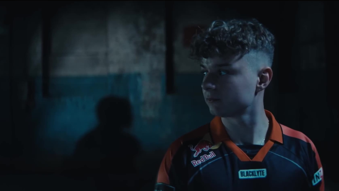 A Valorant player in a Red Bull–branded jersey glances off-frame. The environment is gritty and raw, his expression poised and alert, emphasizing the stakes.