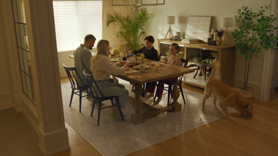 A family gathers around a dining table set for breakfast, with a playful dog entering the scene — all grounded by a Tumble rug that anchors the warmth and energy of the room.