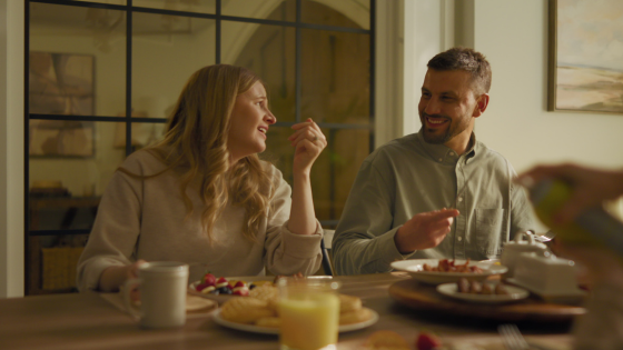Captured in natural light, a couple shares a warm laugh over pancakes and coffee, framed by the inviting tones and texture of the surrounding space.