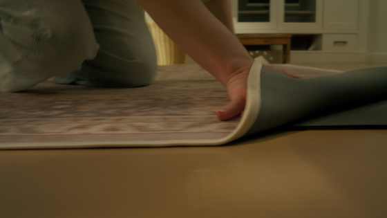 A hand lifts the edge of a Tumble rug to reveal the cushioned underlayer beneath, emphasizing the product’s functional design and easy-to-clean structure.